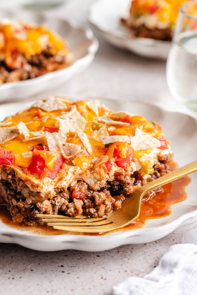 slice of high protein taco casserole. layers of ground venison, greek yogurt, tomatoes, green onion, melted cheese and crushed tortilla chips on a plate with a gold fork.