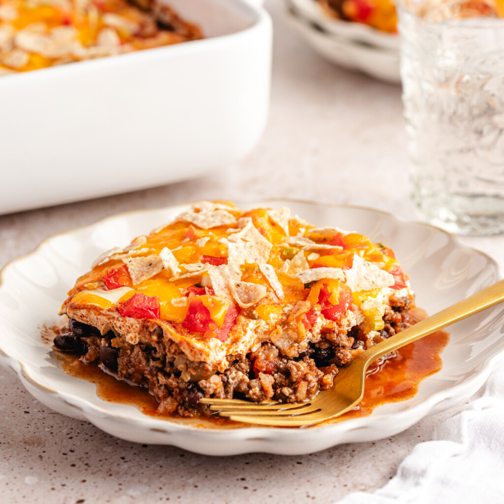 slice of high protein taco casserole. layers of ground venison, greek yogurt, tomatoes, green onion, melted cheese and crushed tortilla chips on a plate with a gold fork. 