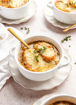 A white bowl of French onion soup garnished with sprigs of thyme on a white plate served with a gold spoon