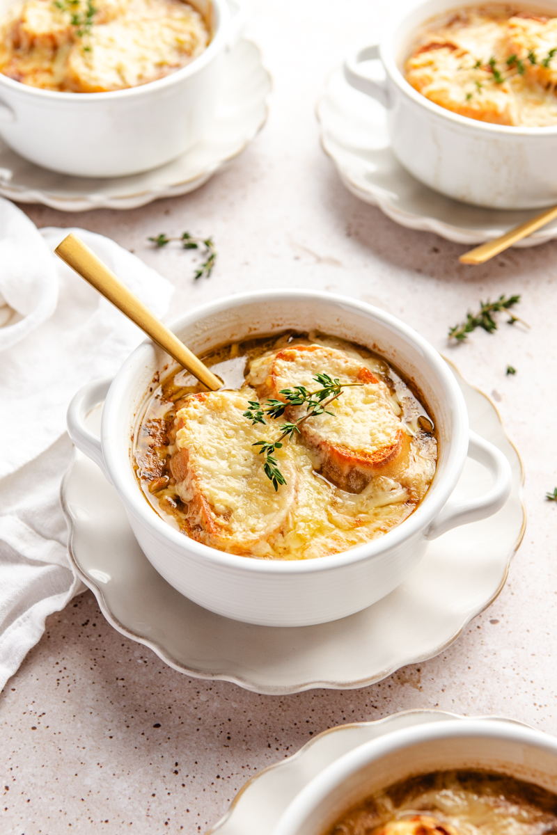A white bowl of French onion soup garnished with sprigs of thyme on a white plate served with a gold spoon