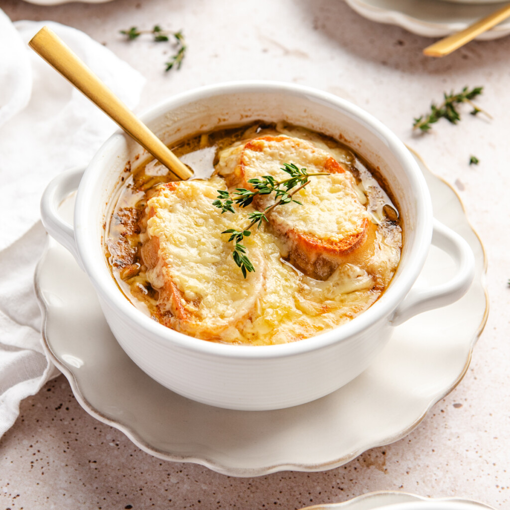 A white bowl of French onion soup garnished with sprigs of thyme on a white plate served with a gold spoon