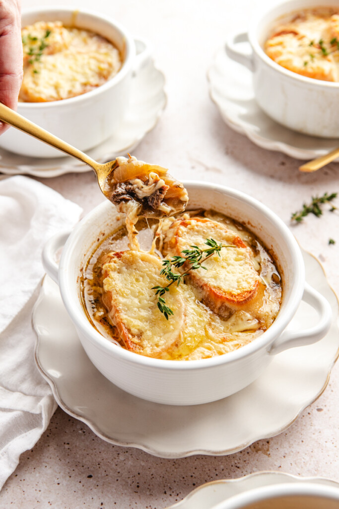 A gold spoon pulling up a cheesy bite of French Onion soup with braised meat from a white bowl