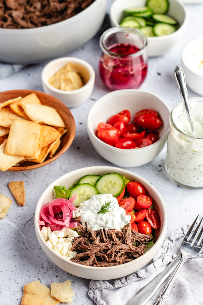 A greek venison power bowl surrounded by bowls of toppings including red onions, tzatziki, pita chips, hummus, cucumber, shredded version and feta cheese