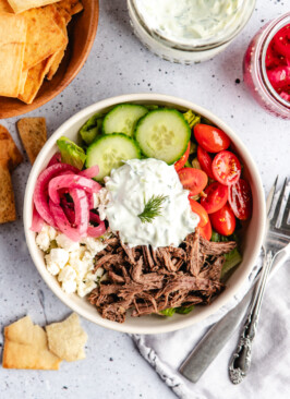 A white bowl with lettuce, cucumbers, cherry tomatoes, red onions, shredded venison, feta cheese and tzatziki sauce on top