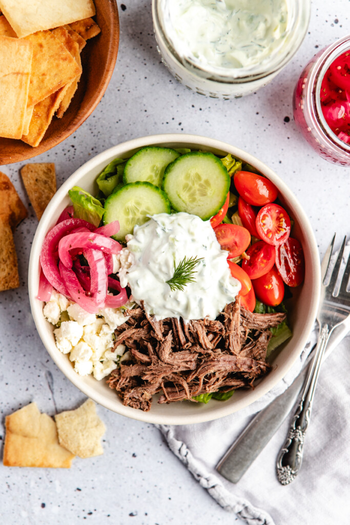 A white bowl with lettuce, cucumbers, cherry tomatoes, red onions, shredded venison, feta cheese and tzatziki sauce on top