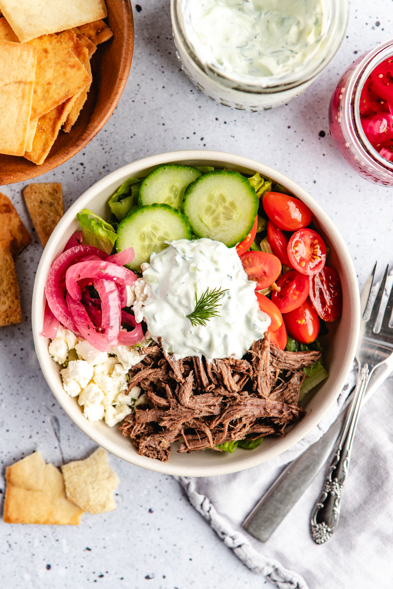 A white bowl with lettuce, cucumbers, cherry tomatoes, red onions, shredded venison, feta cheese and tzatziki sauce on top