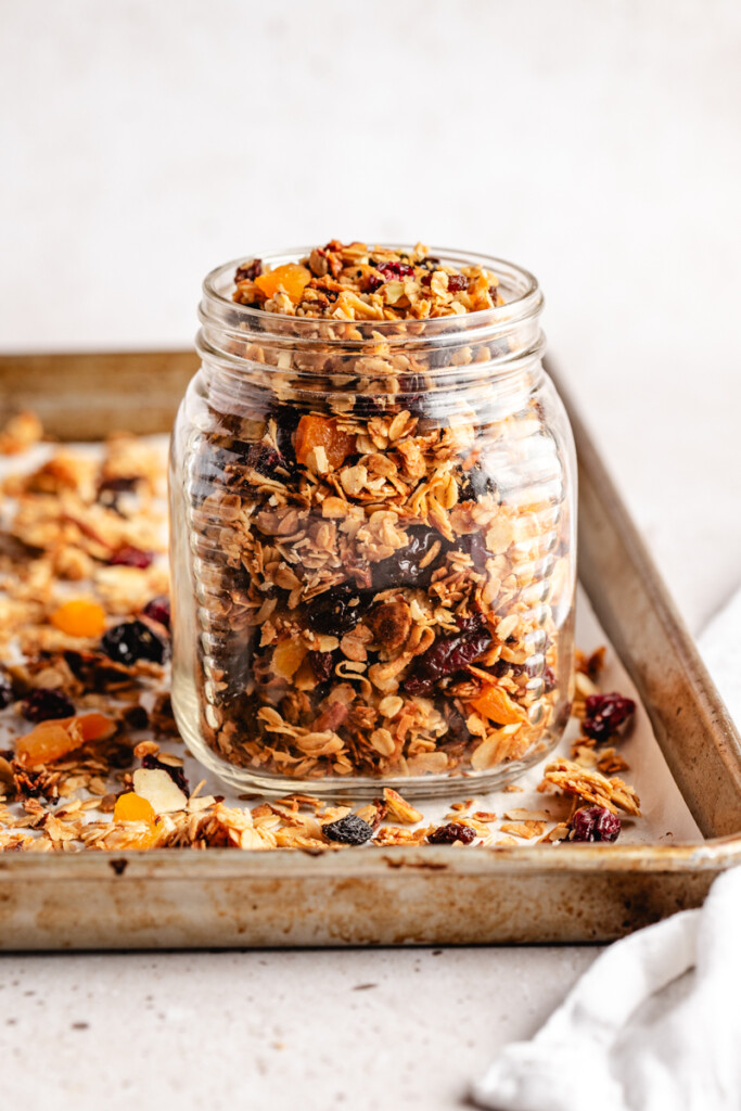 Granola in a glass jar sitting on a baking sheet with granola scattered around