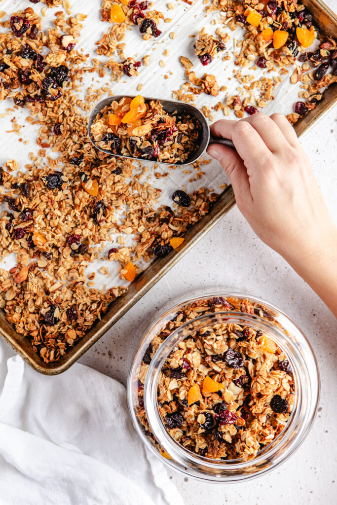 Scooping granola from a baking tray into a glass jar
